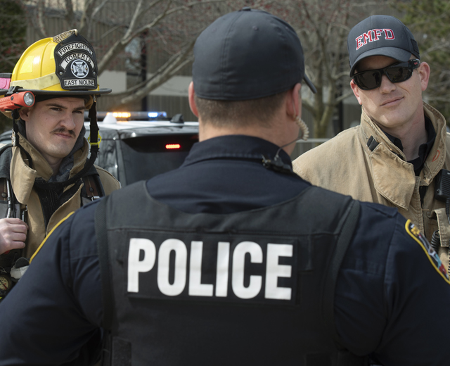 Back of policeman, with "POLICE" in white on his back, facing two firefighters.