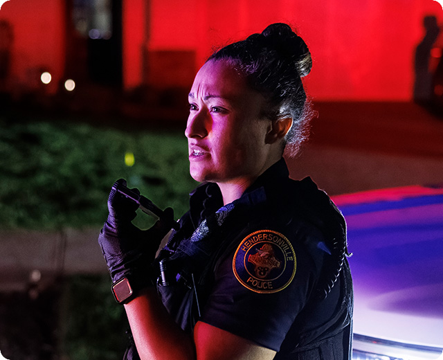 Female law enforcement officer using FirstNet Fusion on a push-to-talk device in front of cruiser 