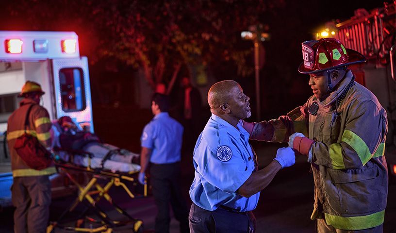 Firefighter and paramedic coordinate near an ambulance at night, while a stretcher with a patient is being loaded inside.