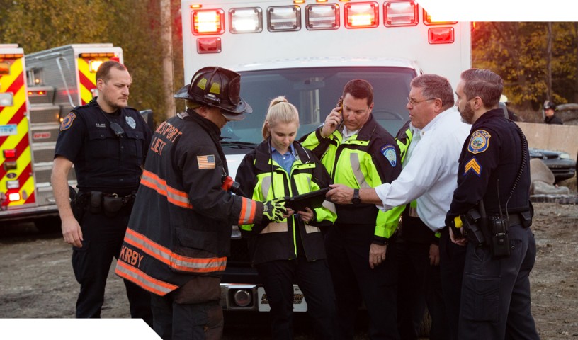 Group of first responders (law enforcement, firefighter, and EMS) gather in front of ambulance and review details on a mobile device.